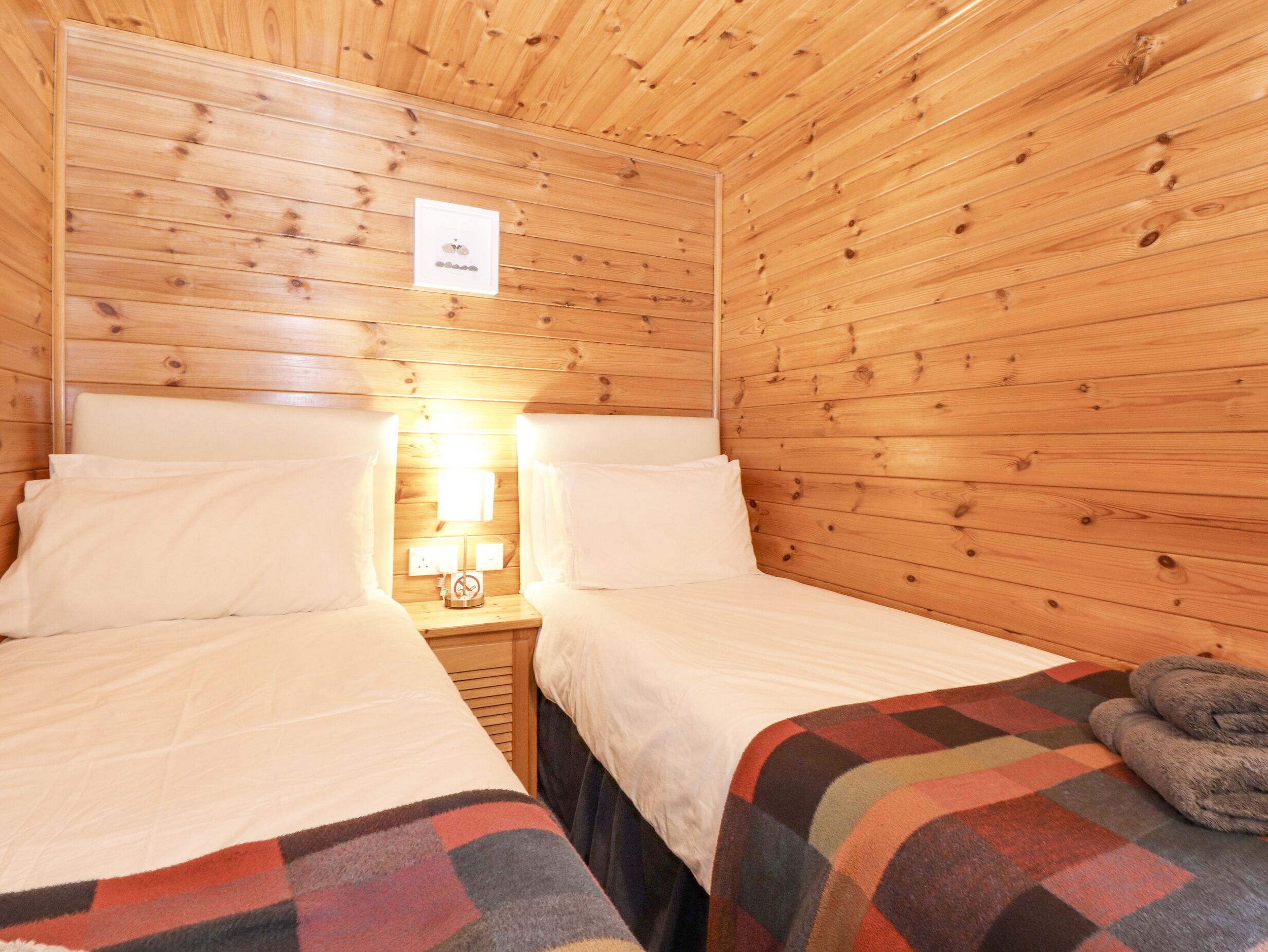 A wooden cabin room with two beds, a nightstand with a lamp, and a blanket on one bed.