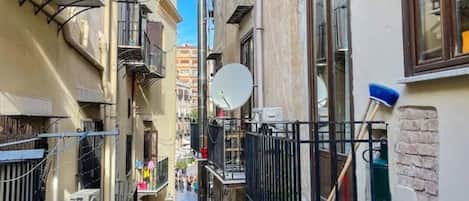 A narrow urban street with buildings on either side, featuring balconies, satellite dishes, and a clear blue sky.