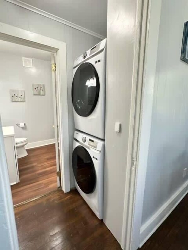 A laundry room with a front-loading washer and dryer, wooden flooring, and a white door leading to a bathroom. - Home Plate Apt 1 - All Star Baseball Rentals (Oneonta)
