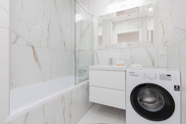 Une salle de bain moderne avec un mur en marbre, une cabine de douche en verre, un meuble-lavabo blanc avec une machine à laver Samsung, et un miroir au-dessus du meuble-lavabo.