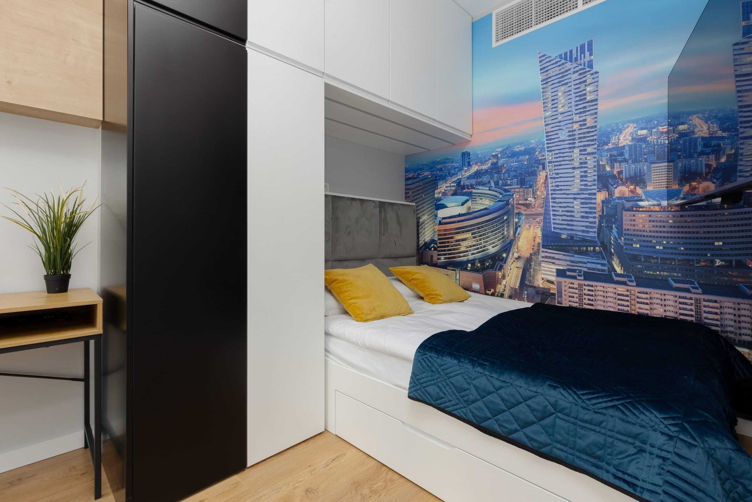 A modern bedroom with a bed, a desk, and a cityscape mural on the wall.