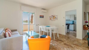 A modern living room with a sofa, a bright orange chair, and a dining table with white chairs. There is a kitchen in the background with a blue and white backsplash. - 3563 Villa del Bacino - App Rosso by Barbarhouse (Porto Cesareo)