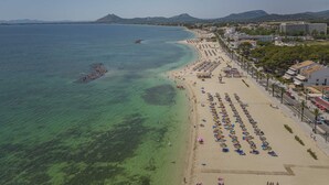 Una spiaggia con file di lettini e ombrelloni, un mare cristallino e edifici sullo sfondo.