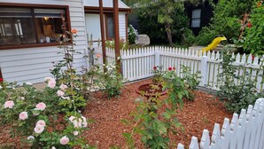 A garden with a white picket fence, various flowering plants, and a bird feeder. - Rose Cottage - Nannup (Nannup)