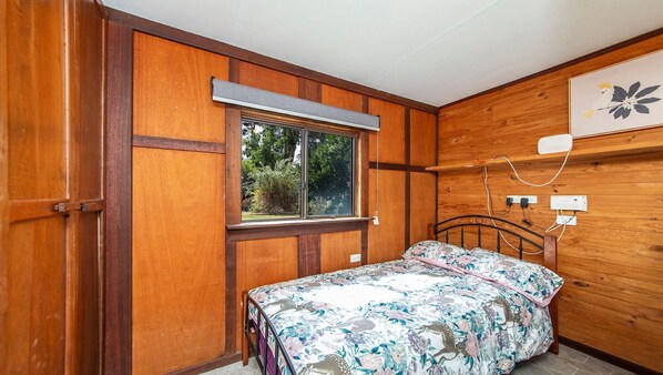 A bedroom with wooden paneling, a bed with a floral bedspread, a window, and a wall-mounted electrical outlet.