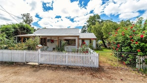 A single-story house with a porch, surrounded by a white picket fence and lush greenery. - Rose Cottage - Nannup (Nannup)