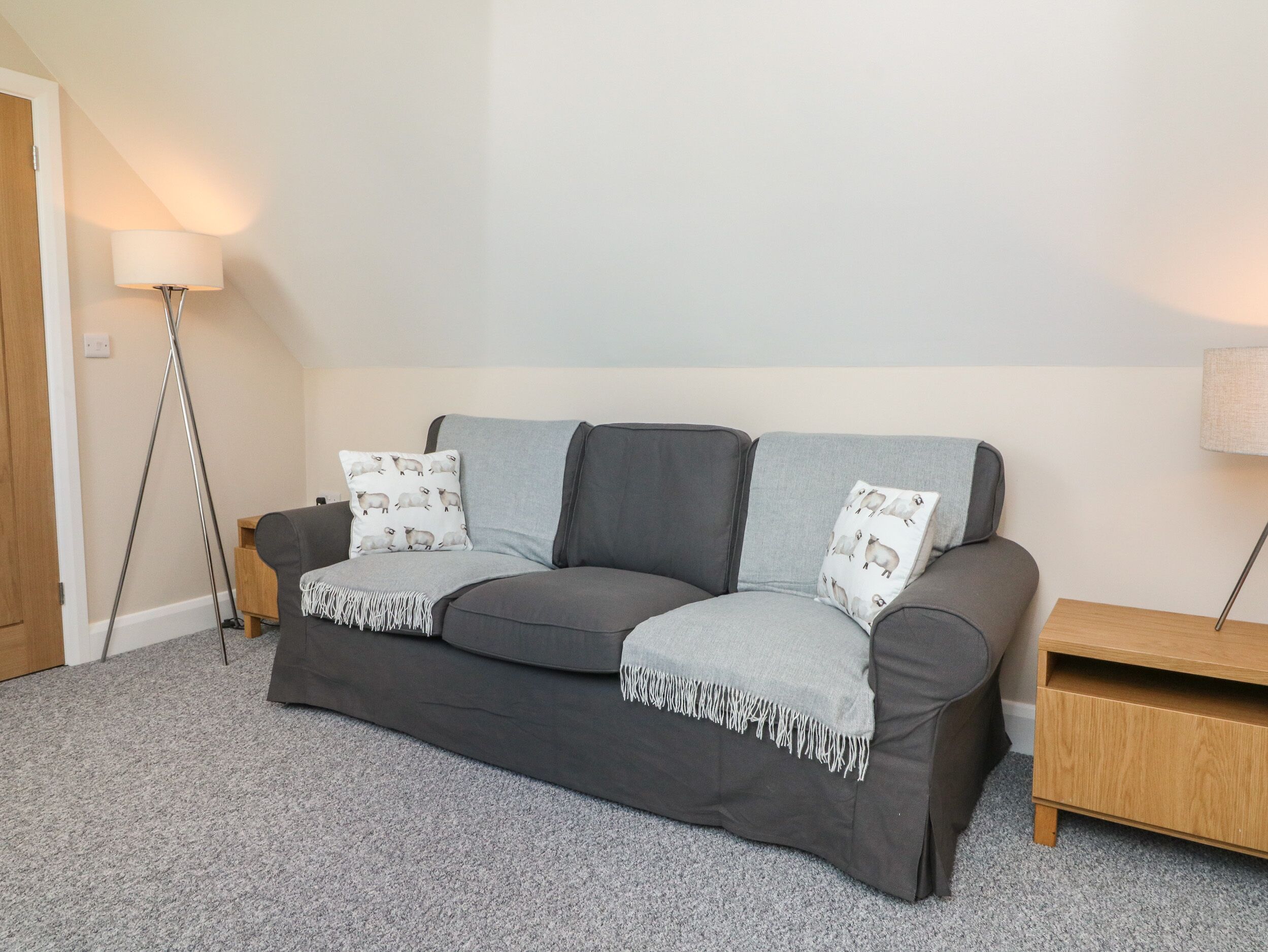 A grey sofa with two pillows, a throw blanket, and a wooden nightstand in a room with a carpeted floor and a lamp.