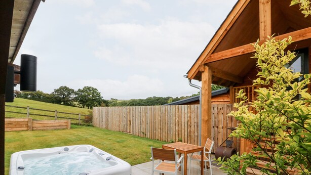 A hot tub in a backyard with a wooden deck, a table and chairs, and a wooden fence. - Spicery Barn Loft (Exeter)