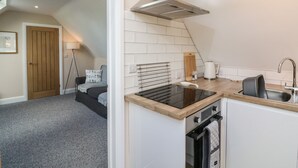 A compact kitchen with a stove, oven, and sink. There is a wooden door and a lamp in the adjacent room. - Spicery Barn Loft (Exeter)