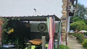 Un edificio rústico con fachada de piedra, un área de estar al aire libre cubierta y un jardín con diversas plantas y flores.