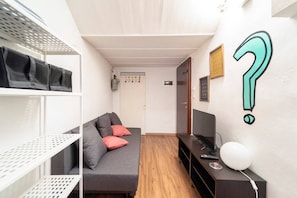 Quarto pequeno com sofá cinza, televisão e prateleira com bolsas.