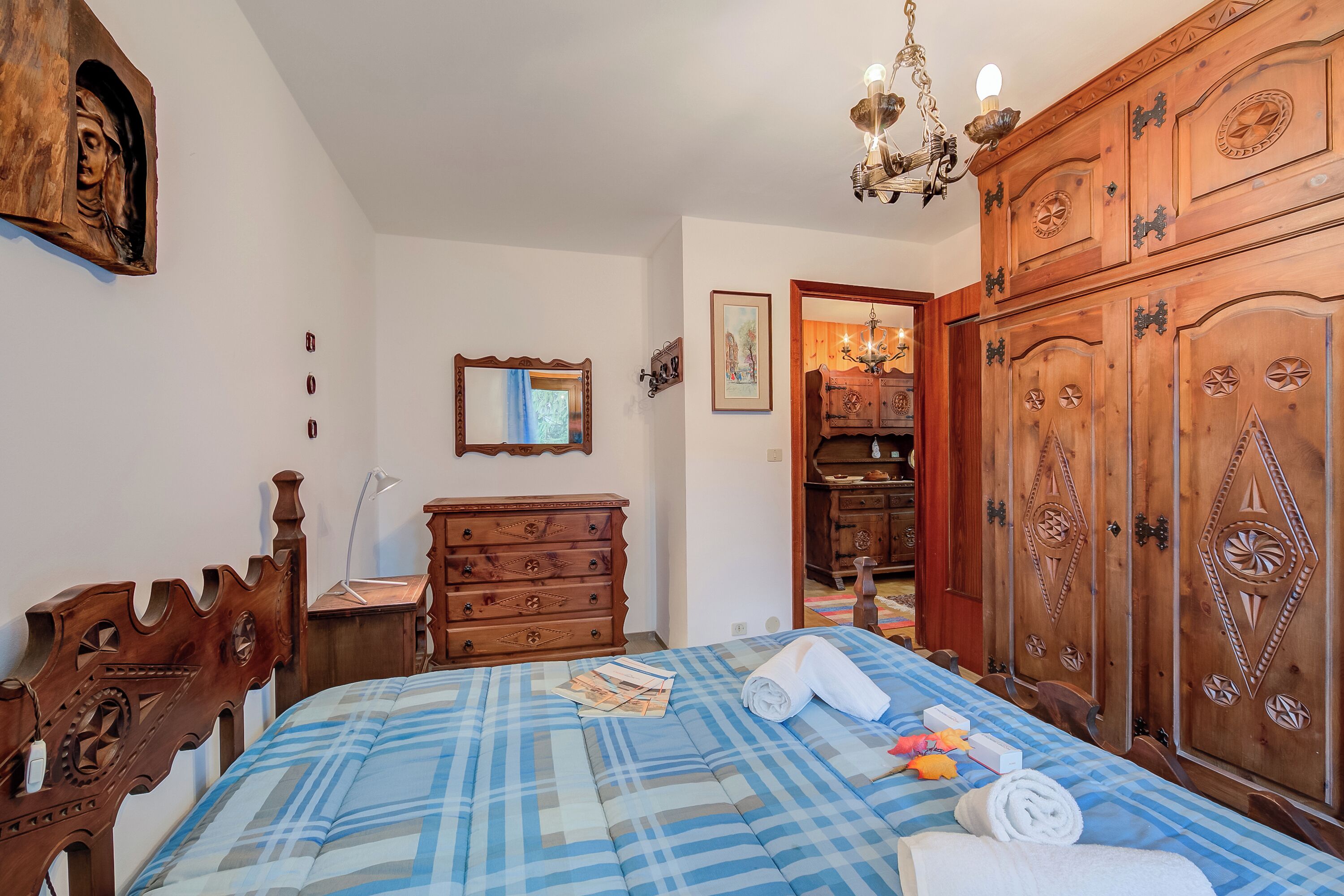 A bedroom with a wooden headboard, a bed with a blue and white checkered bedspread, a wooden wardrobe, and a chandelier.