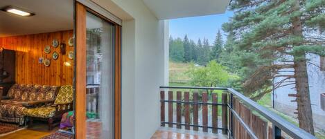 A balcony with a wooden railing, a checkered floor, and a view of a forest.