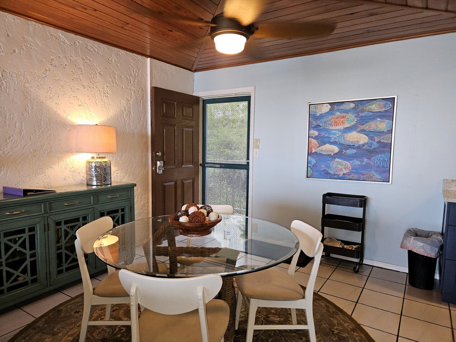 A dining area with a glass table surrounded by white chairs, a wooden ceiling, and a painting of fish on the wall.