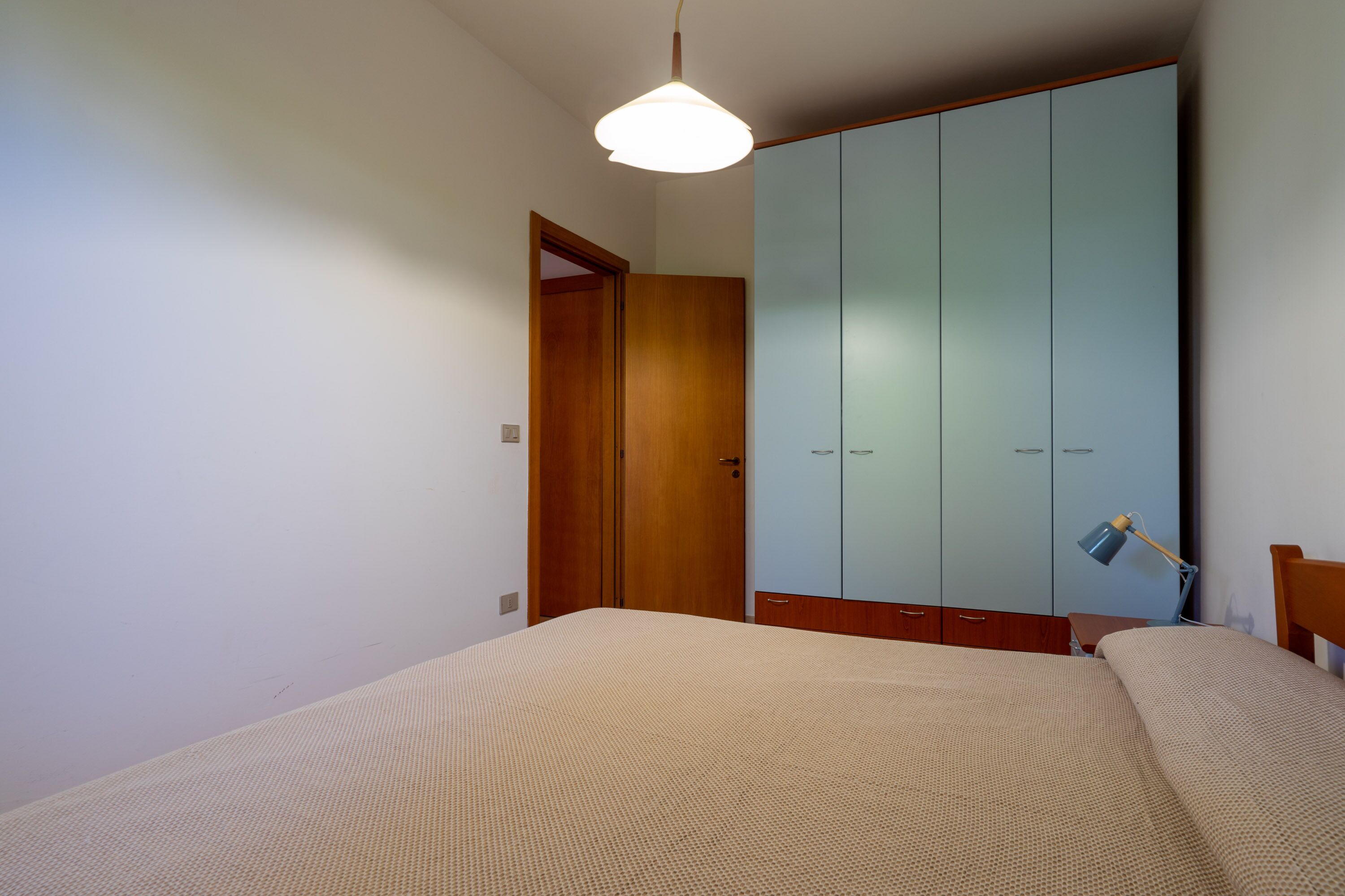 A bedroom with a bed, a wardrobe, a desk with a lamp, and a door.