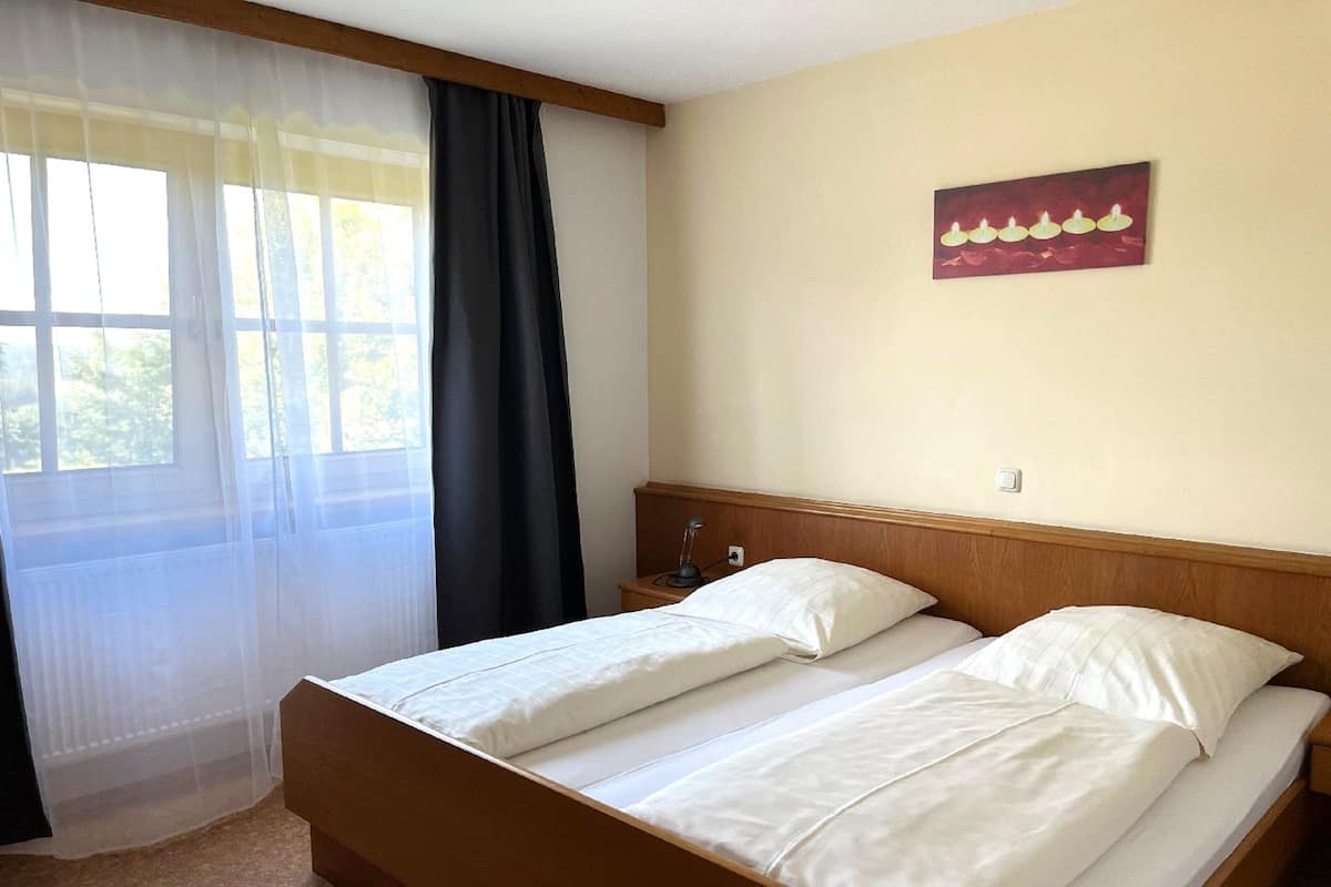 A double bed with a wooden headboard, a bedside table with a lamp, and a window with sheer curtains.