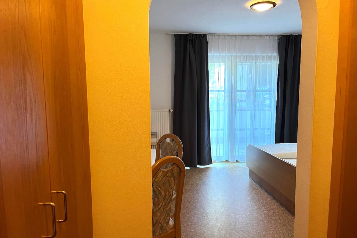 A narrow hallway leading to a bedroom with a bed, a chair, and a window with curtains.