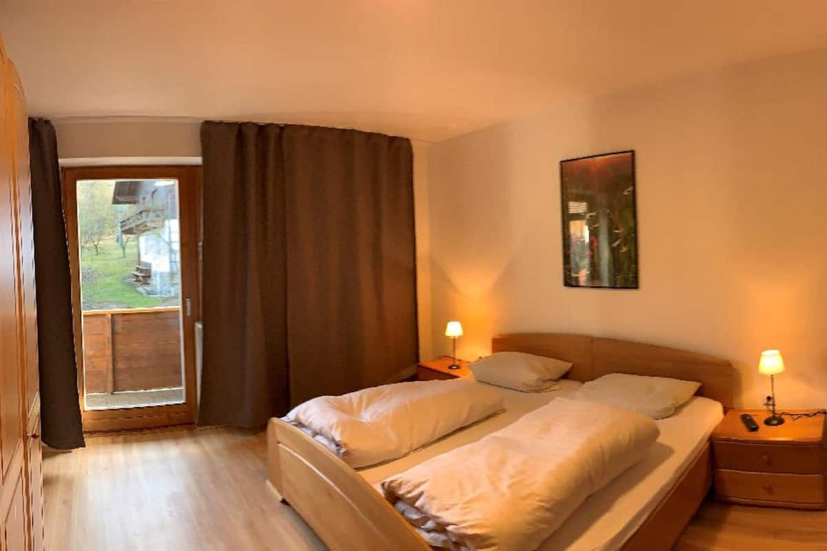 A bedroom with two beds, a wooden floor, a window with curtains, a bedside table with a lamp, and a mirror on the wall.