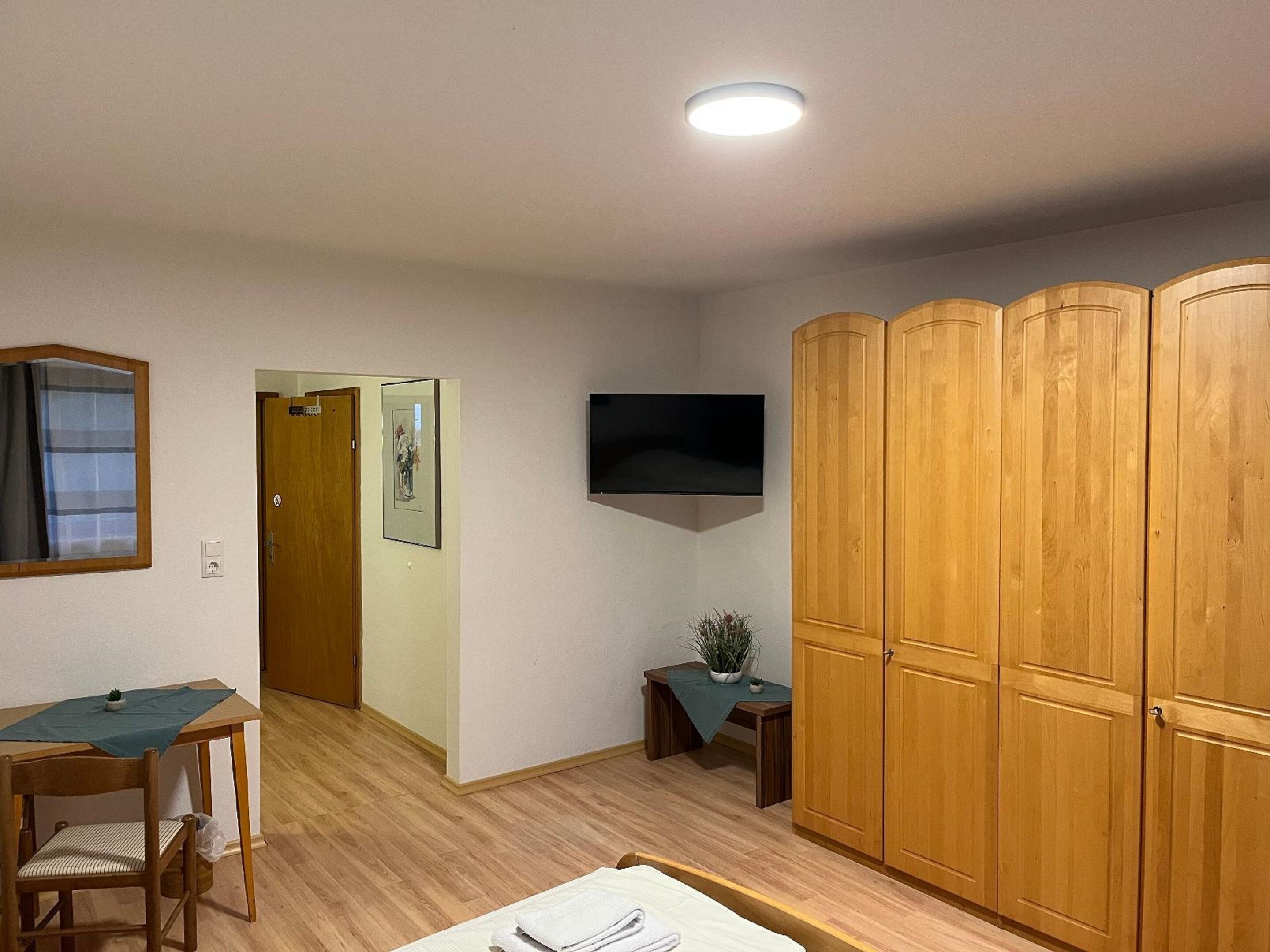 A room with a wooden wardrobe, a flat-screen TV, a small table with a plant, a chair, and a bed.