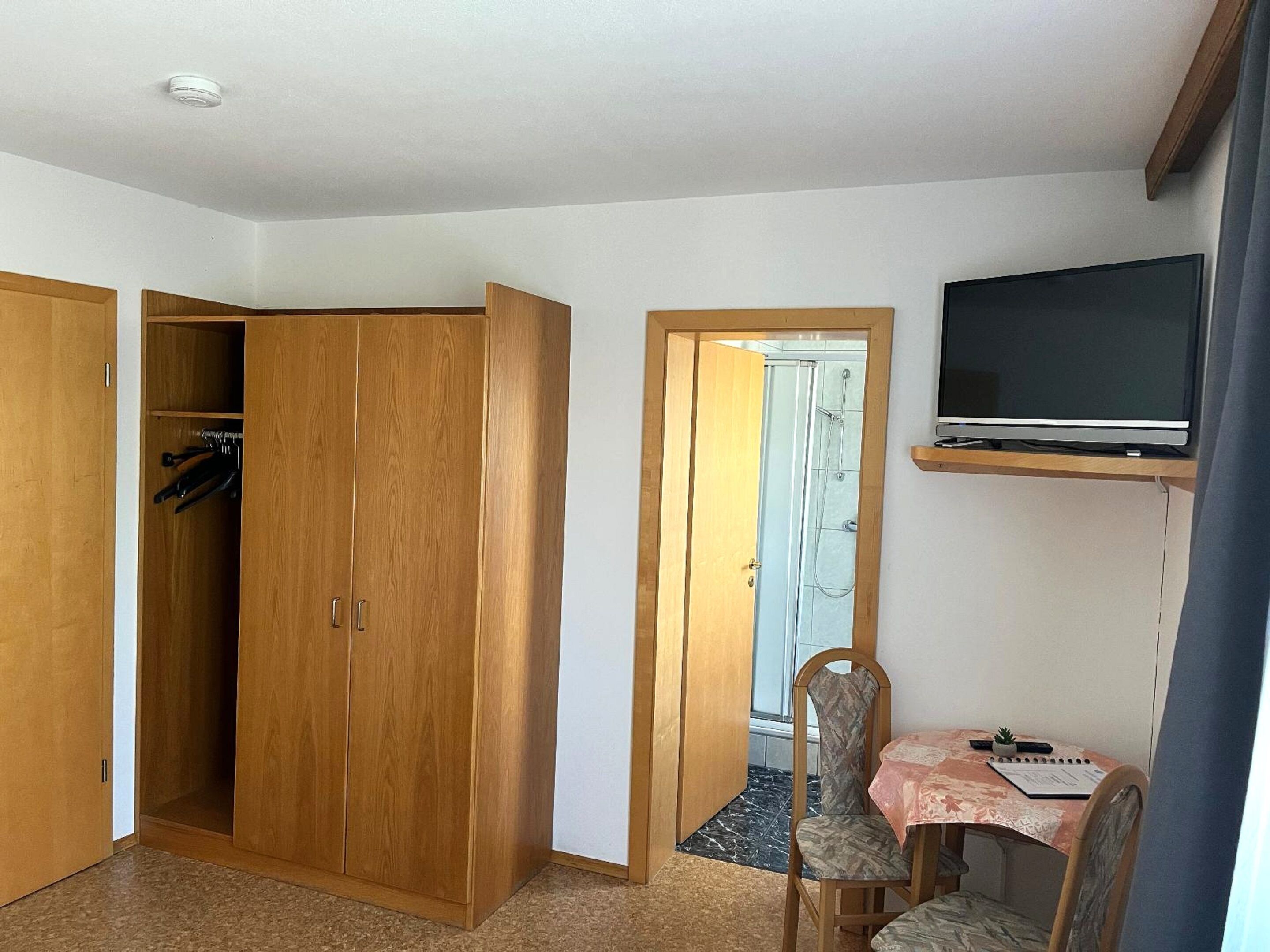 A room with a wooden wardrobe, a shower area, a television mounted on the wall, and a small table with a chair.