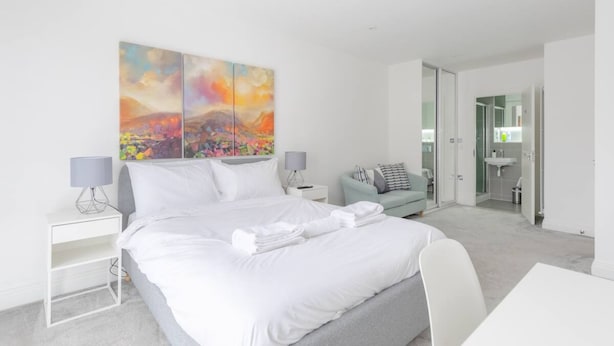 Una habitación de hotel moderna con una cama grande, un escritorio, una silla, un pequeño sofá y un baño visible a través de una puerta abierta.