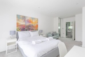 A modern hotel room with a large bed, a desk, a chair, a small sofa, and a bathroom visible through an open door. - Spacious 4BD Townhouse - Near Stratford City! (London)
