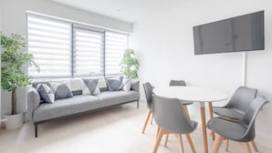 Una sala de estar moderna con un sofá gris, una mesa redonda y sillas grises. Hay dos plantas en macetas y un televisor de pantalla plana montado en la pared.