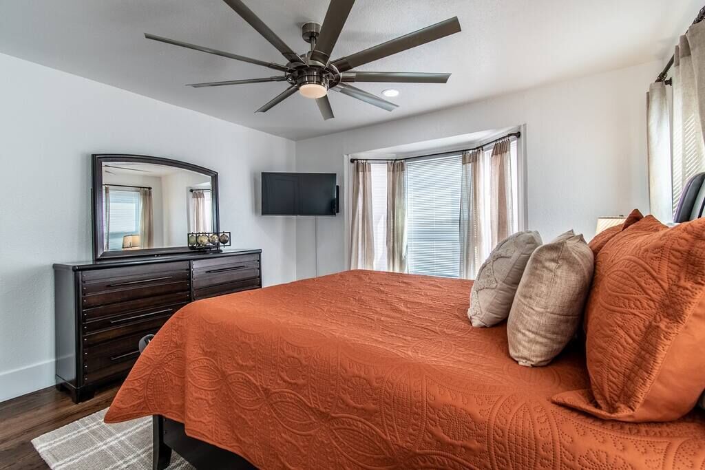 Une chambre à coucher comprenant un lit, un ventilateur de plafond, une commode, un téléviseur et une fenêtre avec des rideaux.
