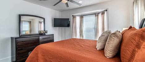 Une chambre à coucher comprenant un lit, un ventilateur de plafond, une commode, un téléviseur et une fenêtre avec des rideaux.