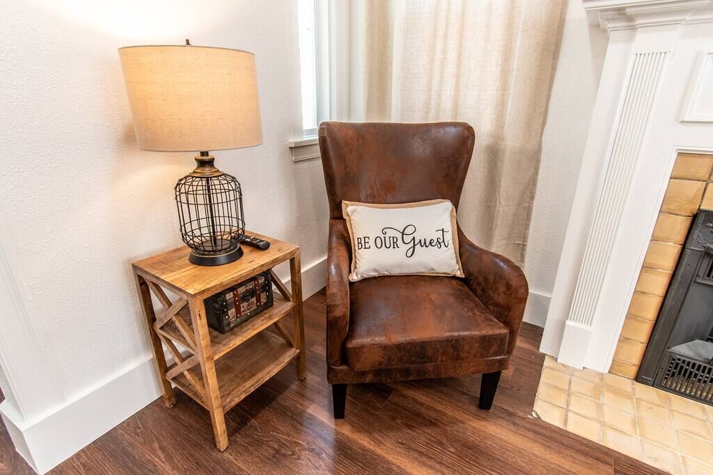 A brown leather armchair with a cushion that reads "BE OUR GUEST" next to a wooden side table with a lamp and a radio.