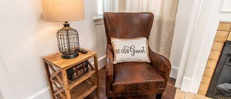 A brown leather armchair with a cushion that reads "BE OUR GUEST" next to a wooden side table with a lamp and a radio.