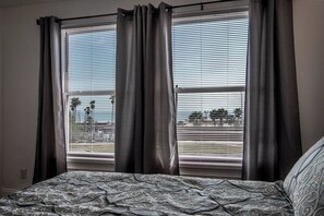A bedroom with a bed, two windows with curtains, and a view of palm trees and a distant landscape.