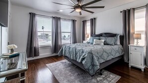 A bedroom with a bed, a ceiling fan, a nightstand, a desk, and a window with curtains.
