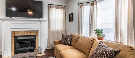 A living room with a fireplace, a sofa, a TV mounted on the wall, and large windows with curtains.