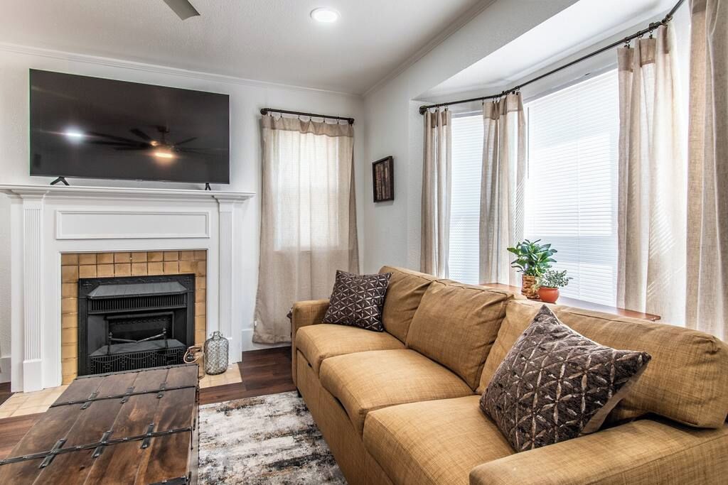 A living room with a fireplace, a sofa, a TV mounted on the wall, and large windows with curtains.
