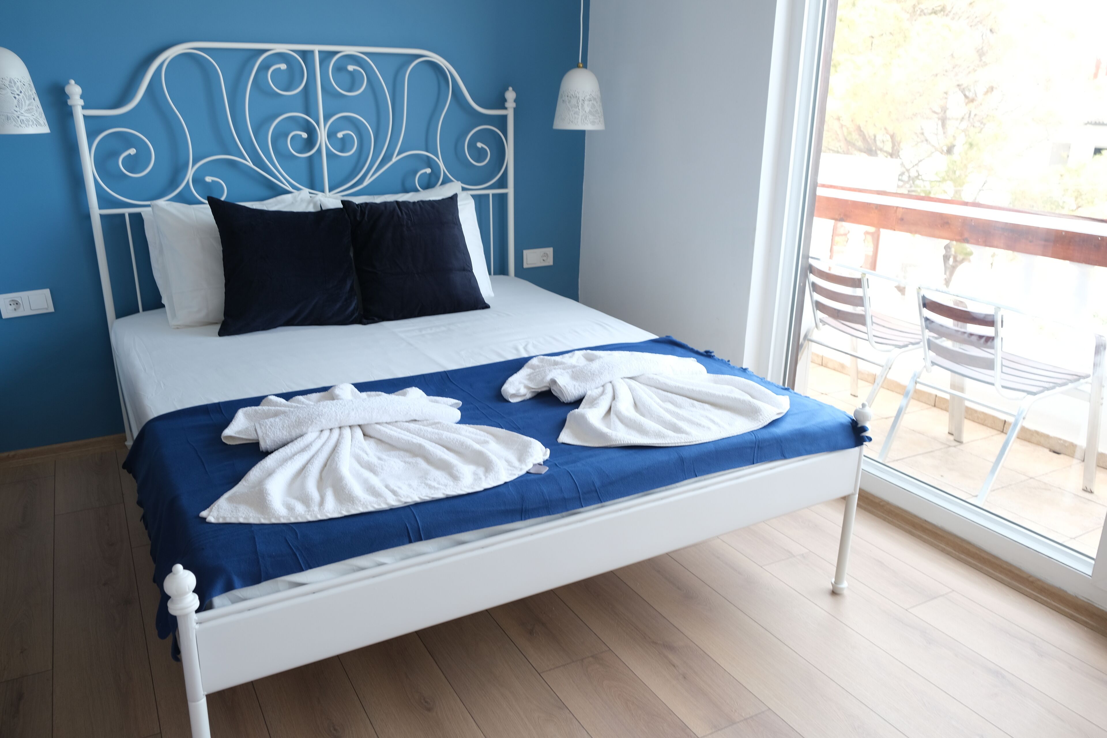 Ein Bett mit weißem Kopfteil und blauer Bettwäsche, zwei weiße Handtücher liegen auf dem Bett, und ein Balkon mit Stühlen.