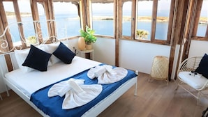 A bedroom with a bed, pillows, a bedside table with a plant, a chair, and a view of the sea. - Arya Hotel Karaburun (Karaburun)