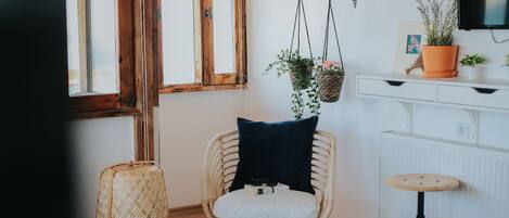 A room with a wicker chair, a small stool, a radiator, and a potted plant on a white dresser.