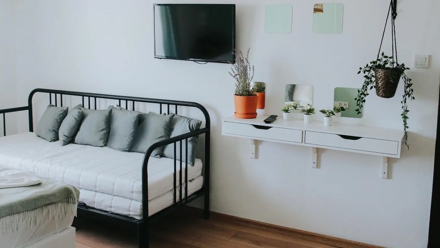 Un dormitorio con estructura de cama metálica negra, ropa de cama blanca, un televisor montado y un aparador blanco con plantas en macetas.