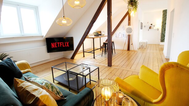 A modern living room with a green sofa, yellow armchair, and a glass coffee table. A Netflix sign is visible on the wall, and there is a staircase leading to another room. - Boho-artstudio: Schicke Designer-wohnung mit Workspace +zentral+wifi+alexa+ (Duisburg)