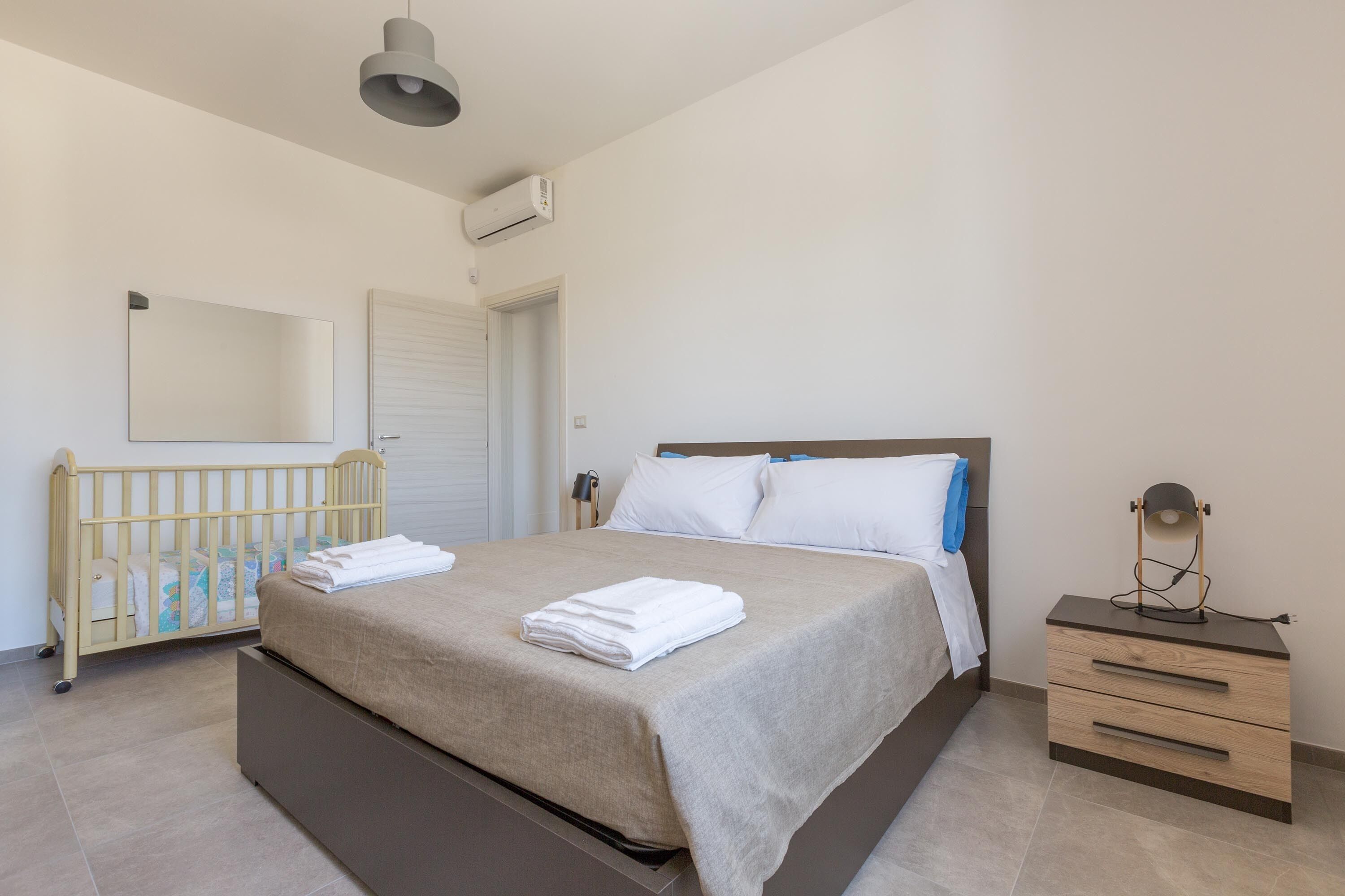 A bedroom with a bed, a nightstand, a crib, a mirror, and an air conditioning unit.