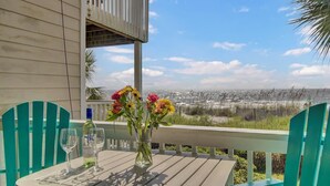 Ein Tisch mit Blumen, zwei Gläsern und einer Flasche auf einer Holzterrasse mit türkisfarbenen Stühlen, mit Blick auf eine Küstenlandschaft.
