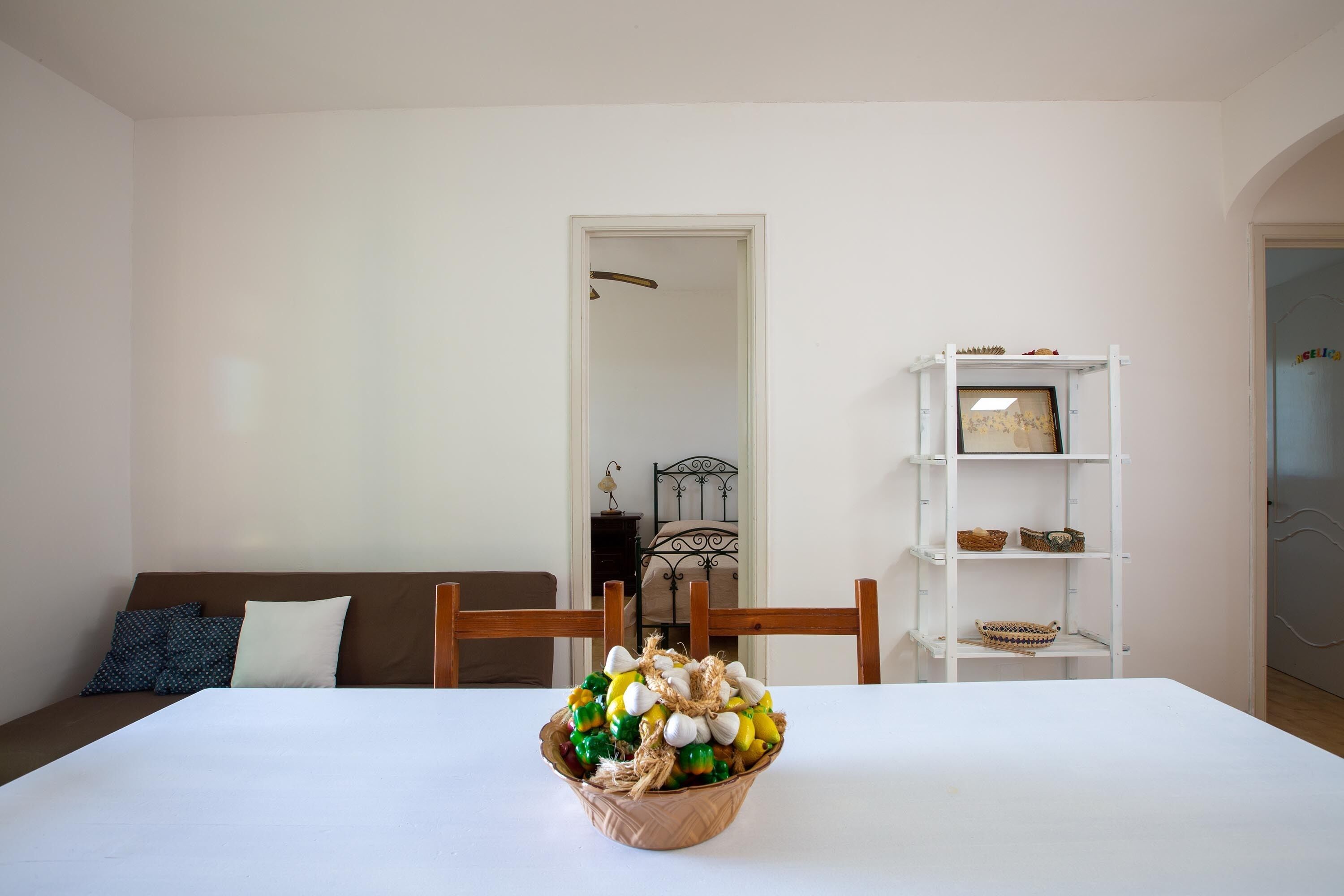 Uma mesa de jantar com uma cesta de objetos decorativos, uma parede branca, uma cadeira de madeira, uma estante branca e uma porta que dá para outro cômodo.