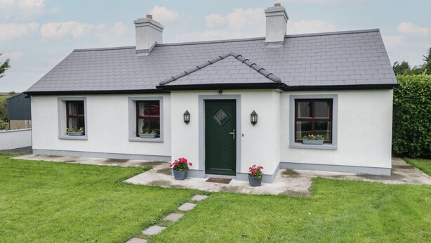 A two-story house with a green door, two chimneys, and a grey tiled roof. There are two windows with flower boxes and a small garden area with stepping stones. - Kate's Cottage (Ballintober)