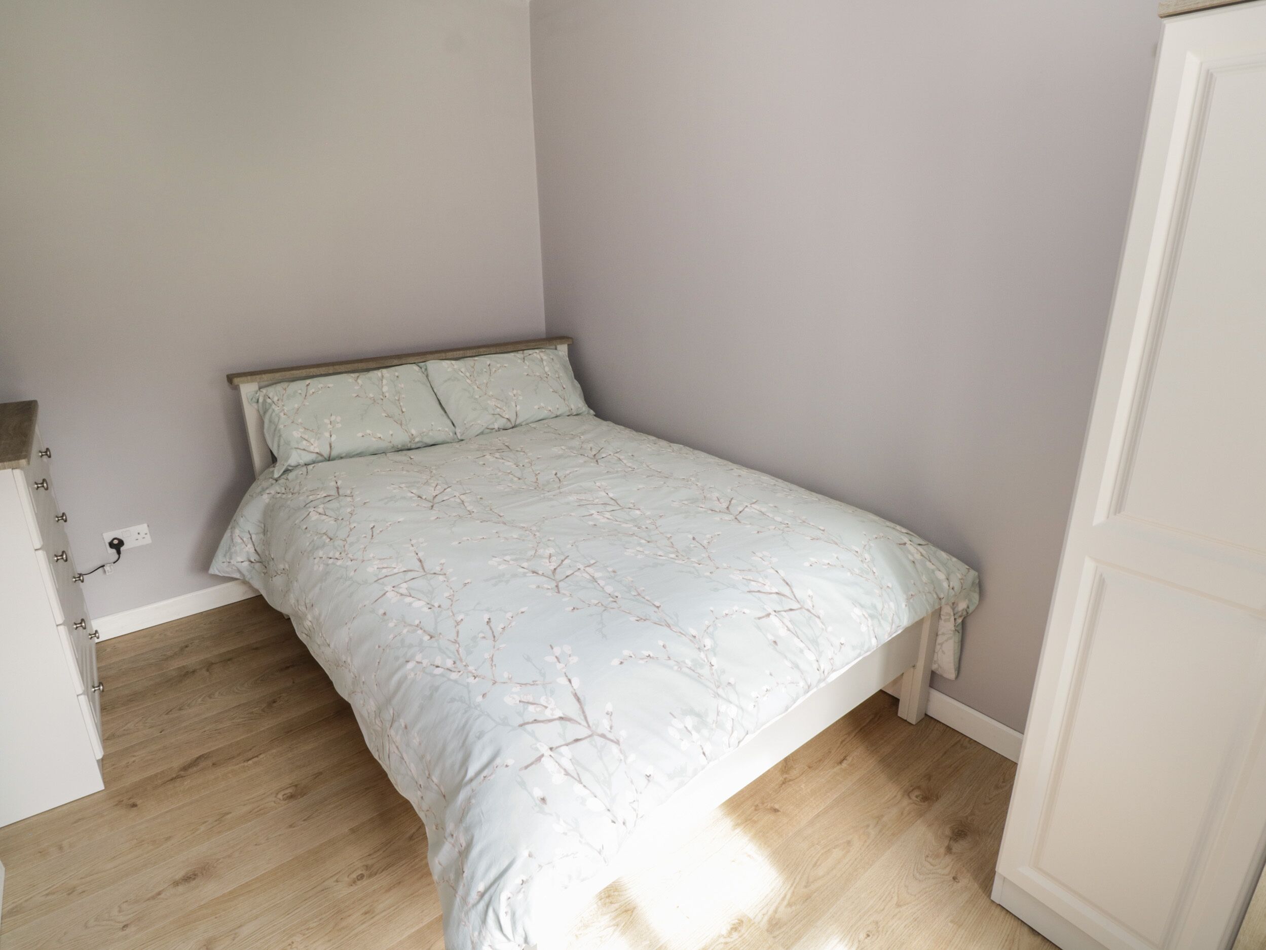 A small, neatly made bed in a room with light-colored walls and wooden flooring.