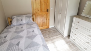 Un dormitorio con puerta de madera, armario blanco, cama con colcha de estampado geométrico y cómoda con espejo.