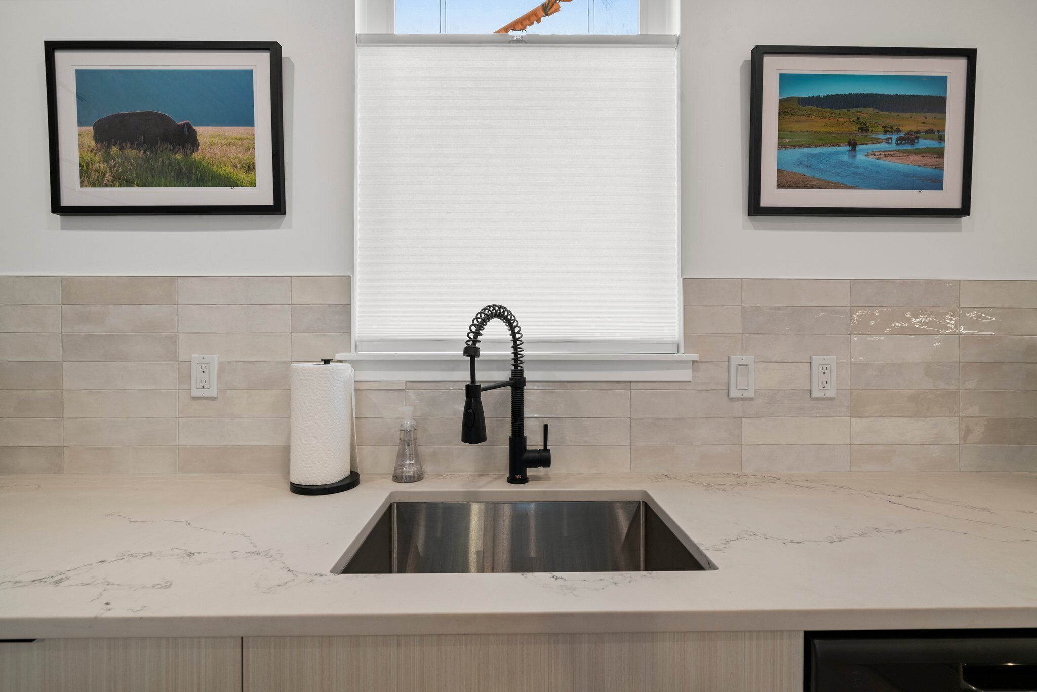 A kitchen with a stainless steel sink, a window with blinds, and two framed pictures on the wall.