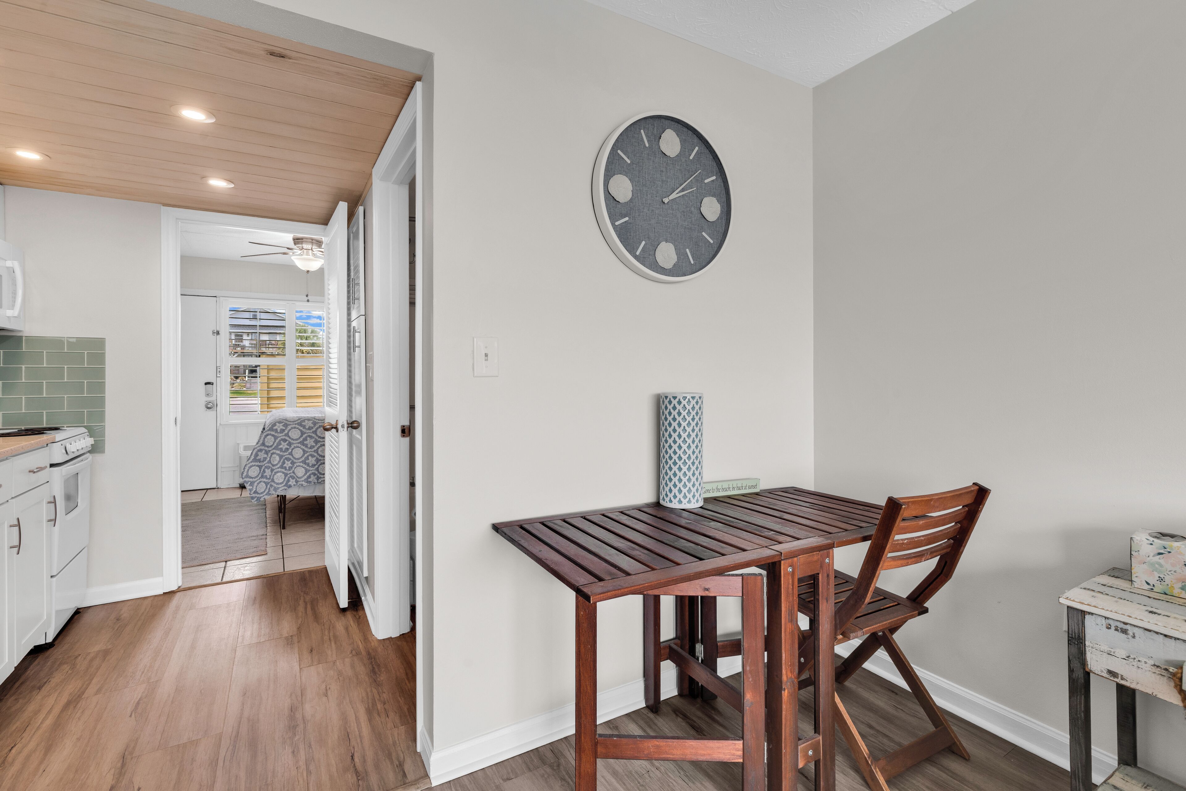A kitchen with wooden cabinets, a dining table with chairs, a wall clock, and a ceiling fan.