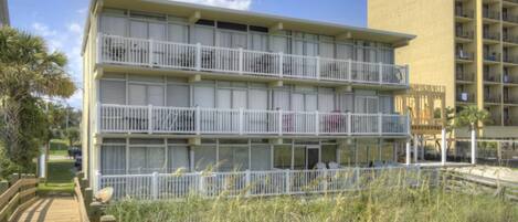 A multi-story hotel with balconies, a wooden boardwalk, and a grassy area in front.