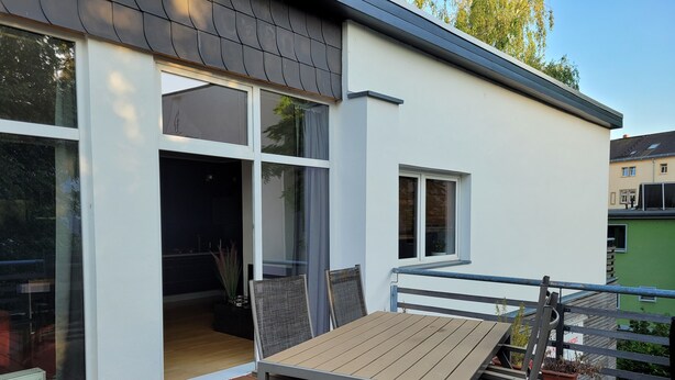 A modern outdoor deck with a wooden table and chairs, a glass door leading to an interior space, and a view of neighboring buildings.